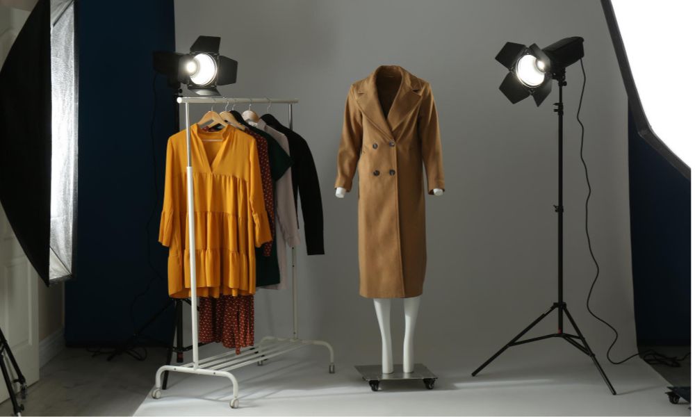 A well-lit photography studio set up for ghost mannequin shots. A light brown trench coat is displayed on a white mannequin, standing next to a clothing rack with an orange dress and other garments. Two large studio lights with softboxes illuminate the scene against a grey background