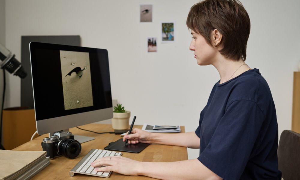 A graphic designer with short brown hair, wearing a dark blue shirt, is using a stylus on a drawing tablet while looking at a computer monitor displaying an image of a bird. An old camera is on the desk next to the monitor