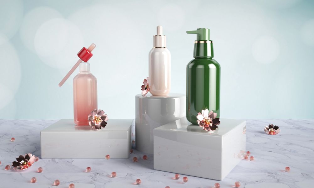 A tall, cylindrical spray bottle with a metallic top and a textured, greenish-grey body stands among several light pink dahlia-like flowers and green leaves, all bathed in dappled sunlight against a light background