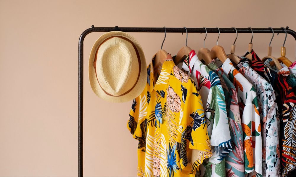 A clothing rack filled with various patterned Hawaiian shirts and a straw fedora hat hanging on the left, set against a plain light brown background