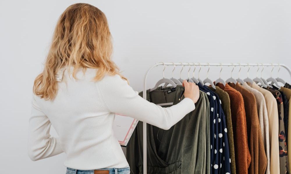 A person with long, wavy blonde hair, seen from behind, stands facing a clothing rack filled with various garments, holding a dark green long-sleeved top on a hanger