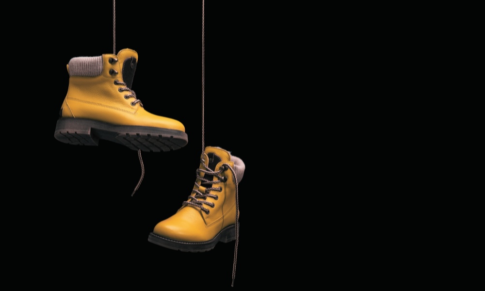 Two yellow work boots with black soles and laces are suspended by strings against a dark background, with one boot higher and slightly to the left of the other