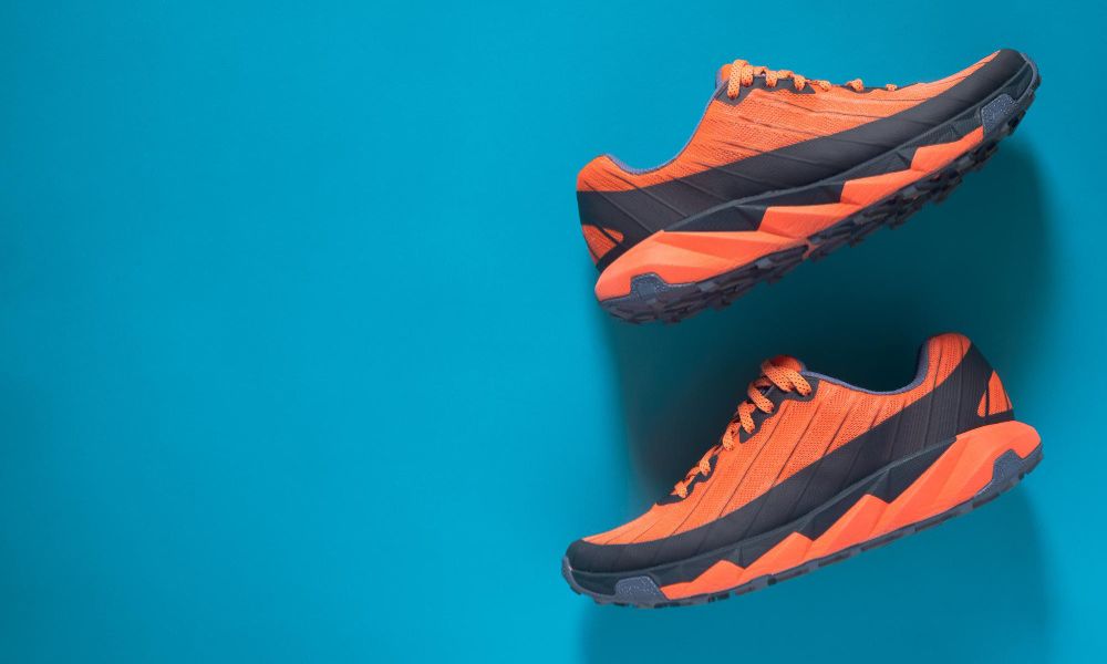 Two vibrant orange and black athletic shoes are arranged diagonally, with the top shoe's toe pointing right and the bottom shoe's toe pointing left, against a bright blue background