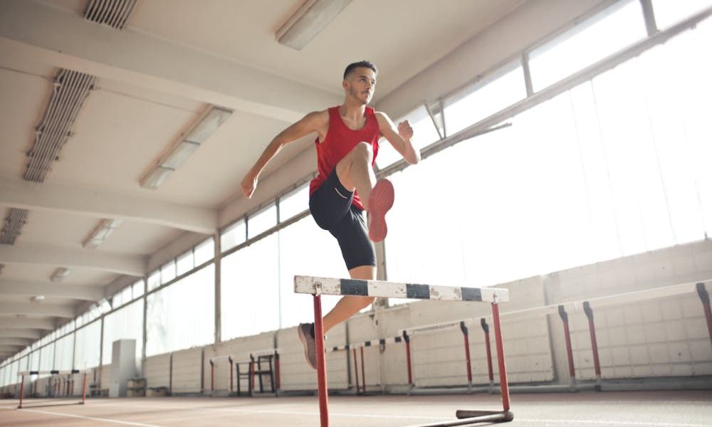A male athlete in a red tank top and black shorts is mid-air, jumping over a hurdle on a track. The indoor sports arena has large windows and bright lighting