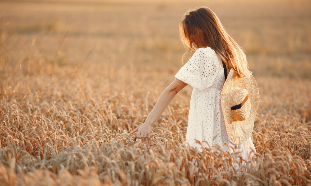 A female with long, flowing brown hair, wearing a white eyelet dress and carrying a straw hat on her back, gently touches the golden wheat stalks in a sunlit field, looking downwards