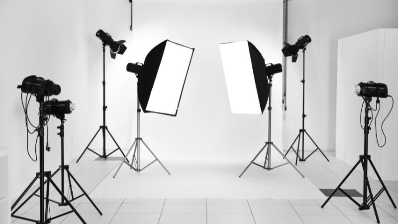 A professional photography studio with a white seamless background and tiled floor is equipped with multiple studio lights on stands, including softboxes and spotlights, arranged for a product shoot