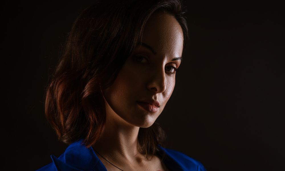Low-key portrait of a woman with short brown hair wearing a blue top, dramatically lit from one side against a dark background