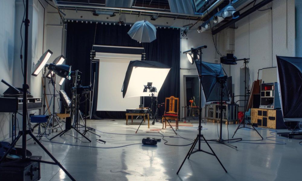 A spacious and modern empty photo studio with a white floor, grey walls, and a black curtain backdrop. Various professional lighting equipment, including softboxes and umbrellas, are set up around the room, along with a white screen and a wooden chair