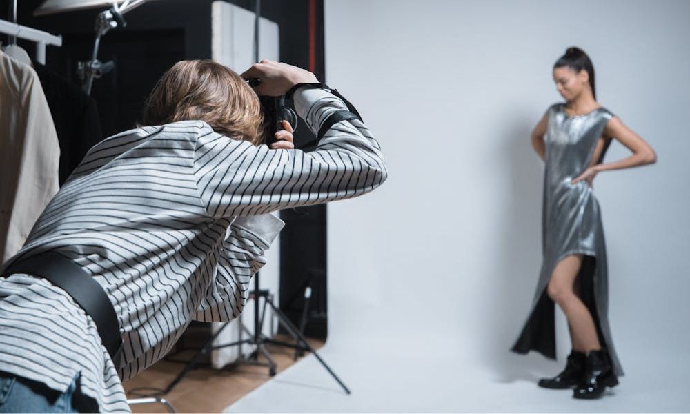 A blonde photographer, seen from behind, is kneeling and taking a photo of a female model striking a pose in a silver, sleeveless dress with a side slit, against a plain white studio backdrop