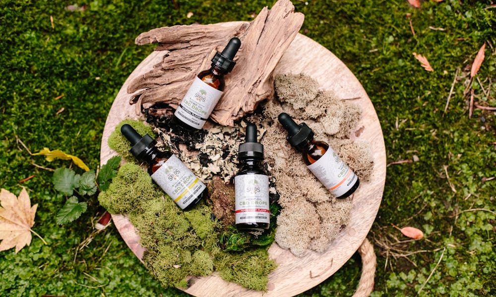 A close-up, high-angle flat lay of four dropper bottles of "SEEDS CBD DROPS" on a round wooden tray, surrounded by various types of moss and a piece of driftwood.