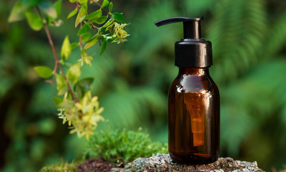 A packshot of an amber glass bottle with a black pump dispenser, a skincare product, placed on a mossy rock. Green leaves and a blurred natural background are visible, suggesting an outdoor setting