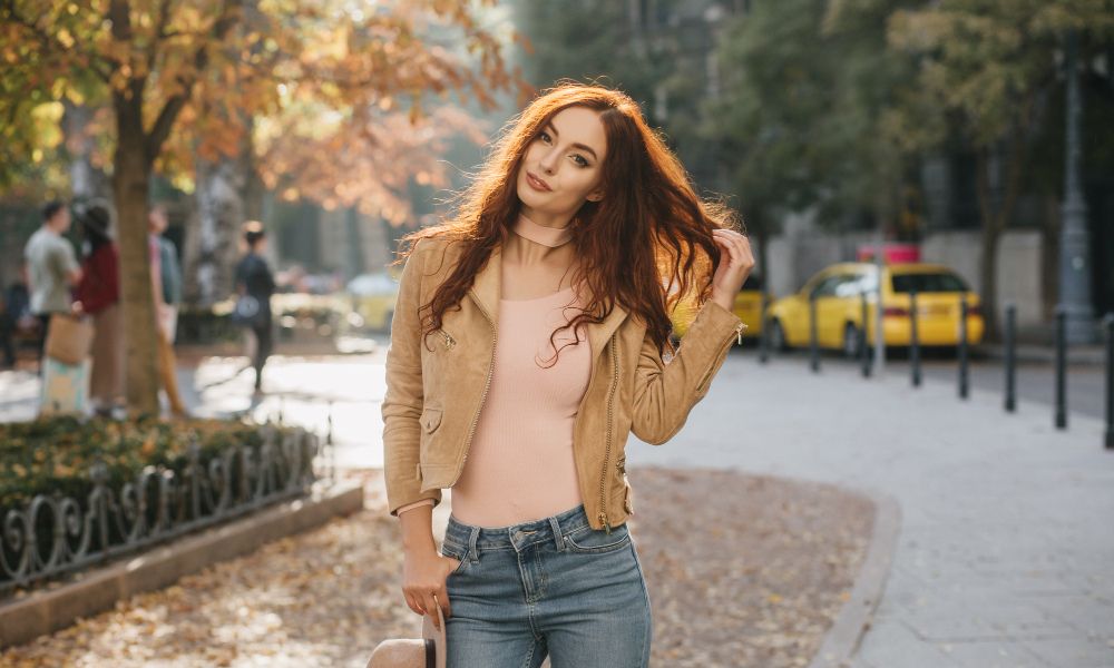 An outdoor portrait of a young woman with long, wavy red hair, wearing a beige jacket, pink top, and blue jeans, posing on a tree-lined street with autumn leaves, and holding a hat in one hand