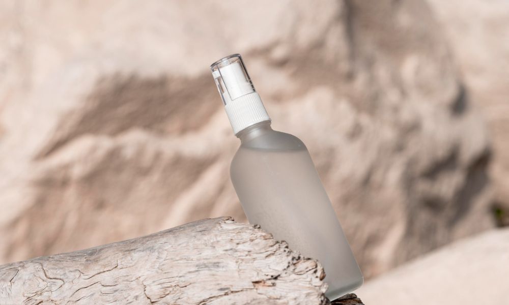 A translucent spray bottle with a white cap is angled on a piece of light-colored driftwood, set against a blurred background of a sandy or rocky natural environment