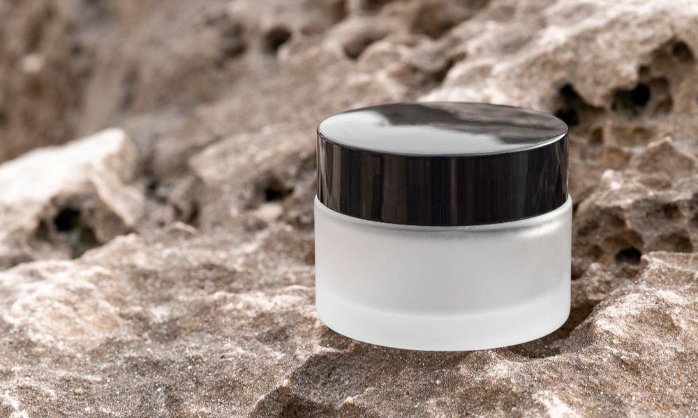 A white, cylindrical cosmetic jar with a black lid sits on a textured, light brown rock surface. The background is a blurred rocky terrain