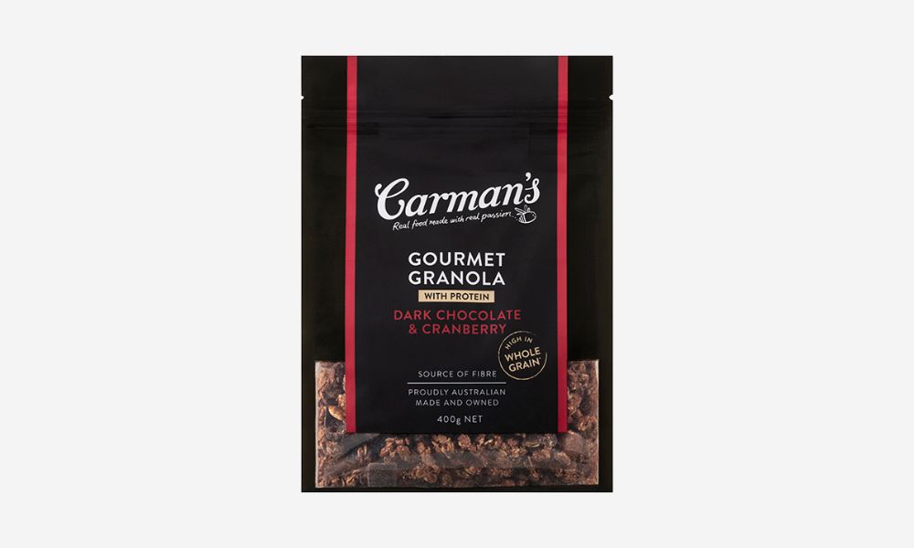 A dark pouch of Carman's Gourmet Granola with Dark Chocolate & Cranberry, showing "WITH PROTEIN" and "HIGH IN WHOLE GRAIN" labels