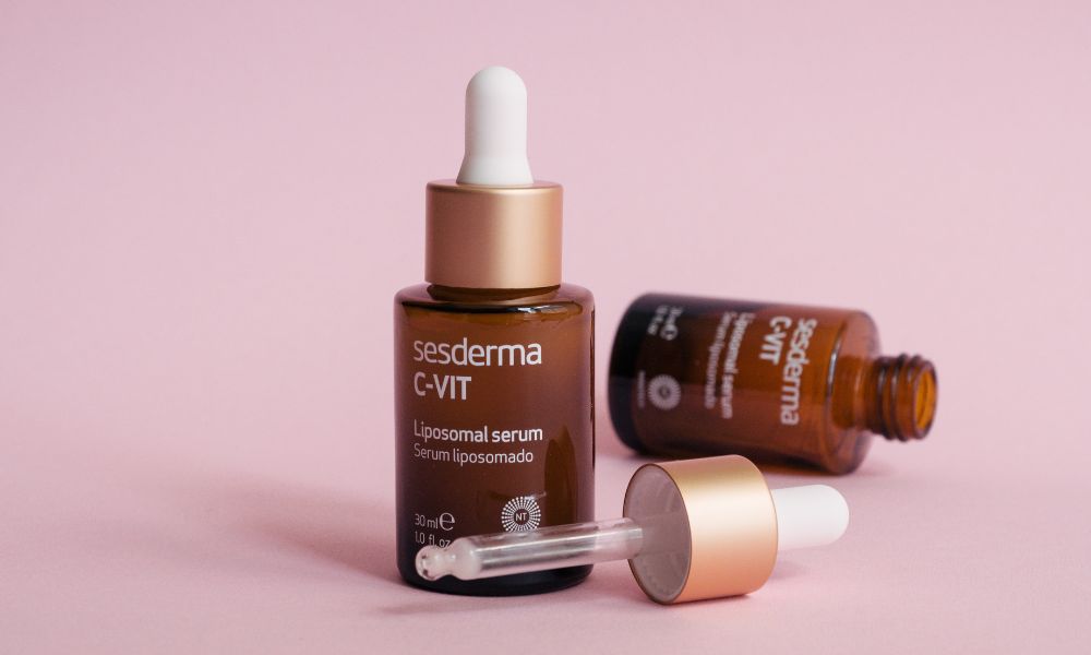Two amber glass dropper bottles of Sesderma C-VIT Liposomal serum on a pink background. One bottle is standing upright with its dropper in place, while the other is lying on its side with the dropper detached