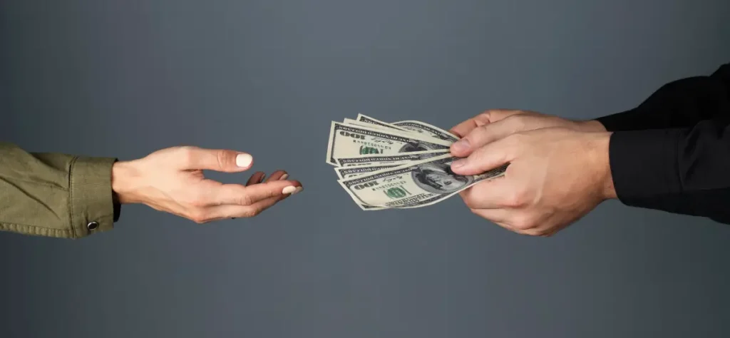 Two hands exchange money against a gray background; one hand on the left is open and ready to receive, while the hand on the right offers several one hundred dollar bills