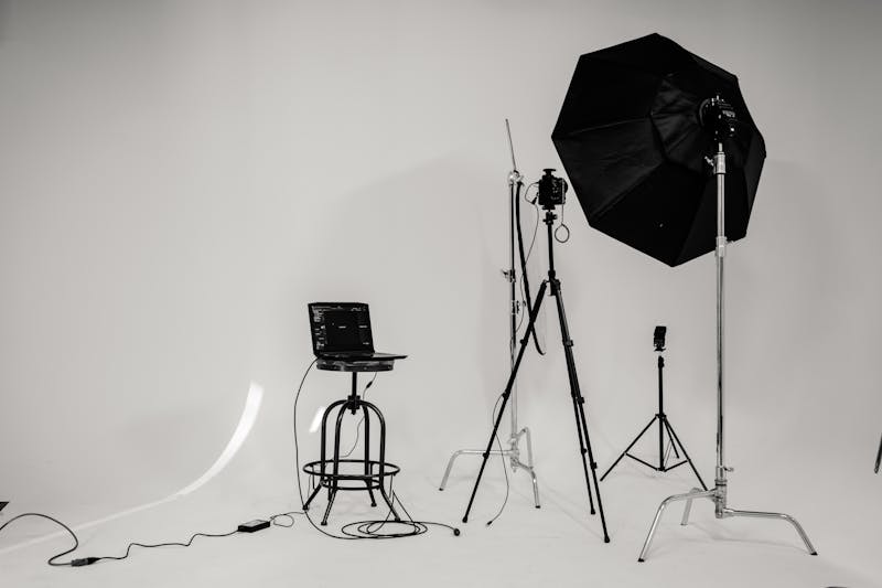 Minimalist photo studio setup with a laptop on a stool, camera on a tripod, large softbox lighting, and various photography equipment arranged on a white background
