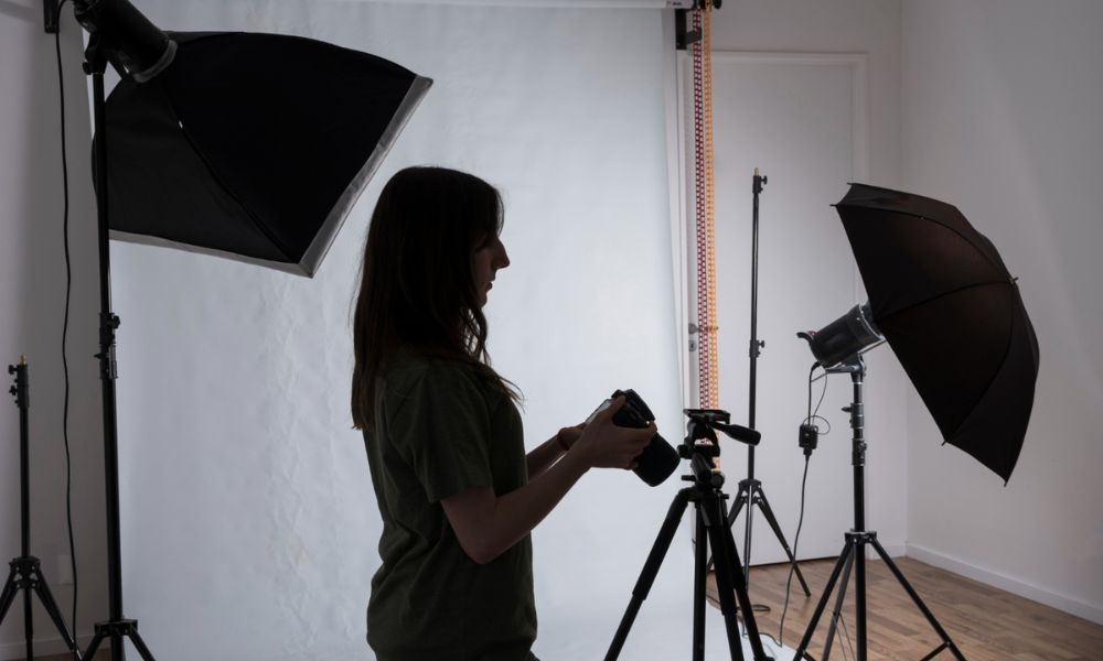 A dark silhouette of a female photographer stands in a studio, holding a camera on a tripod, surrounded by various lighting equipment including softboxes and umbrellas, against a white and grey background