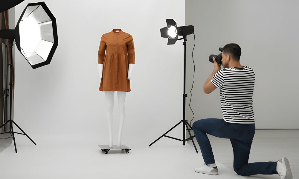 A male photographer in a striped shirt and jeans is kneeling to take a picture of a brown dress on a white mannequin in a well-lit studio. Two large studio lights are also visible