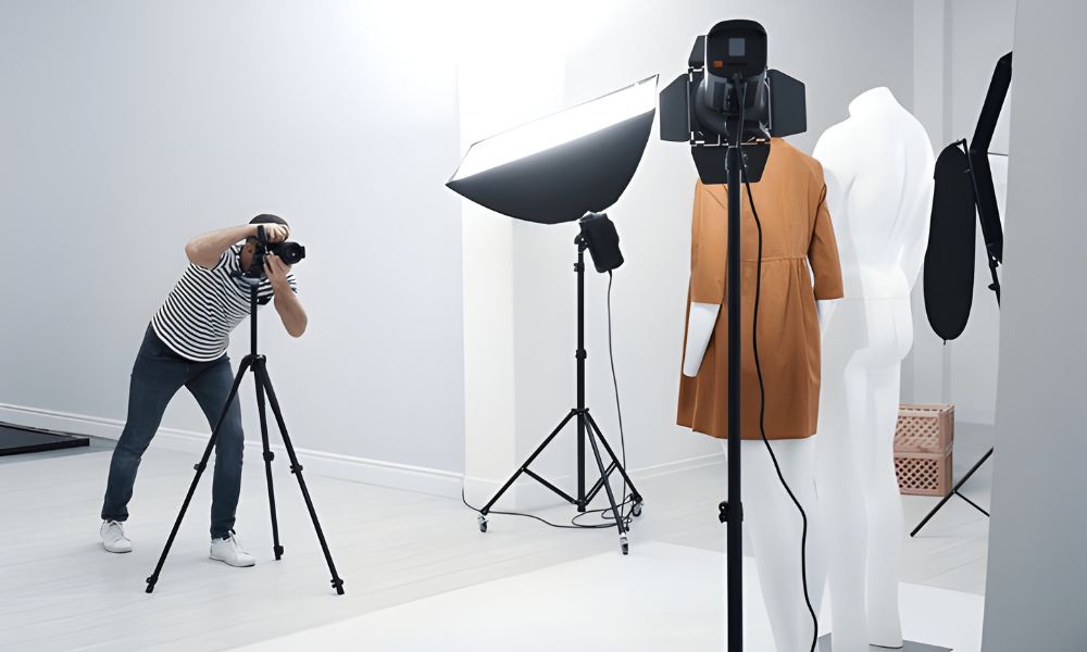 A photographer in a striped shirt and jeans is kneeling to take a picture of a brown dress on a white mannequin in a well-lit studio. Another white mannequin stands to the right, and studio lighting equipment is also visible