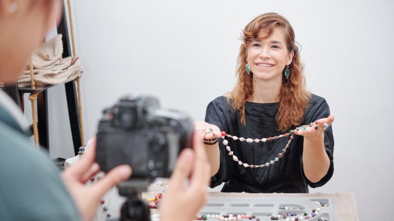 A photographer, seen from behind with a camera on a tripod, is taking a picture of a smiling woman with wavy brown hair who is holding up two necklaces made of colorful beads