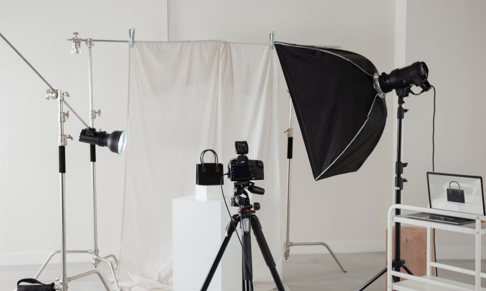 A professional photography studio setup features a camera on a tripod, aiming at a small black handbag placed on a white pedestal, surrounded by various lighting equipment and a white backdrop, with a laptop showing the shot on the right