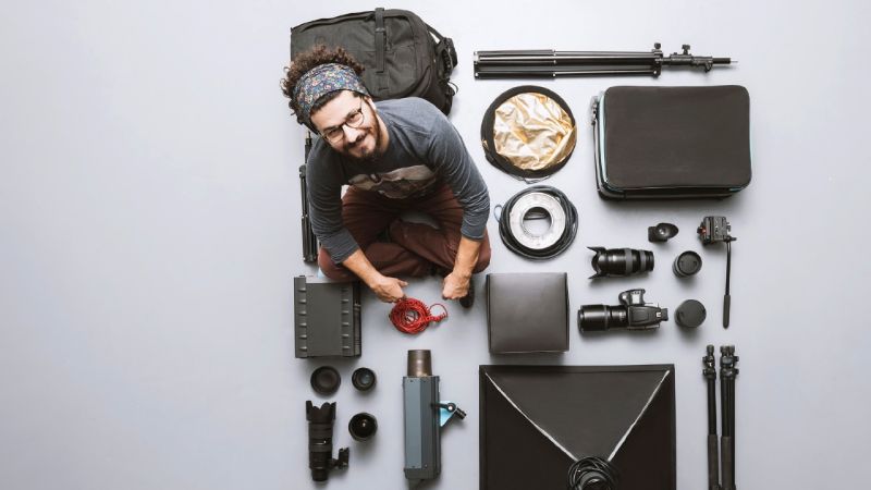 Male photographer sitting on the floor surrounded by neatly arranged photography equipment including cameras, lenses, tripods, light reflectors, cables, and camera bags