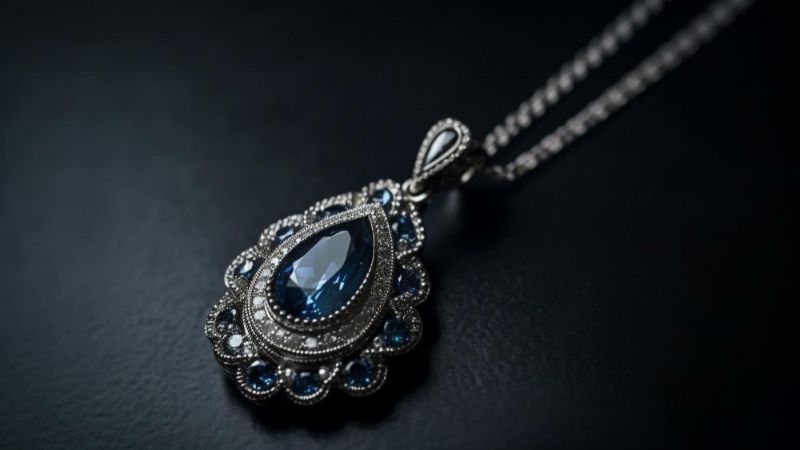 A close-up shot of an elegant silver necklace with a large, teardrop-shaped blue gemstone surrounded by smaller blue and clear stones, resting on a dark, textured surface