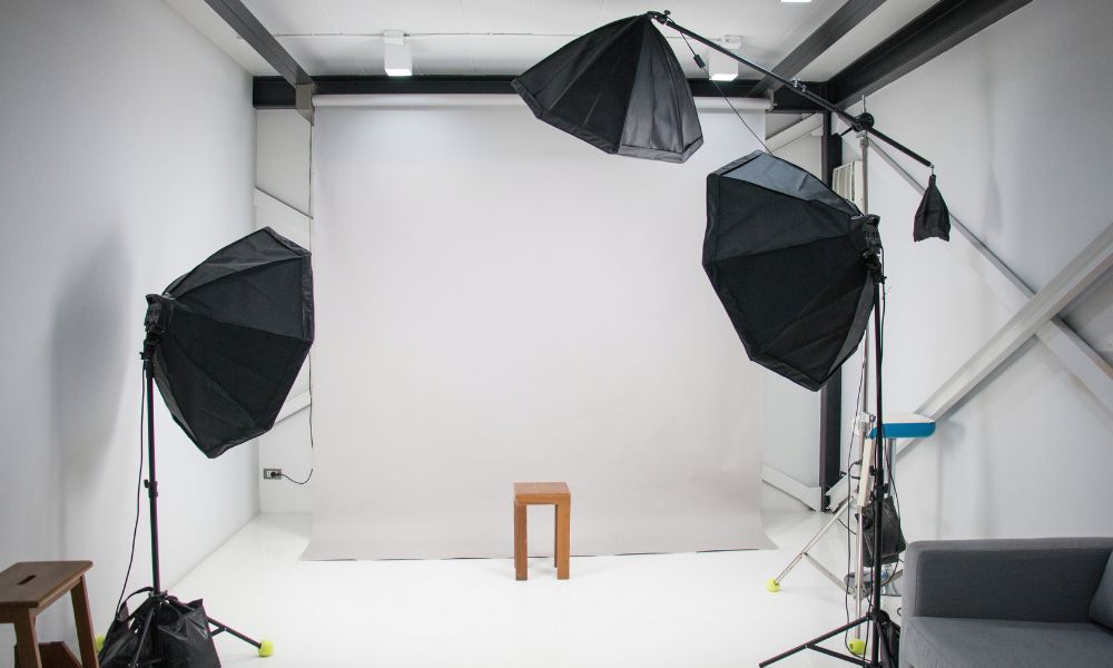 A professional photography studio with a seamless white paper backdrop unfurled, a wooden stool in the center, and three black studio lights with large softboxes set up around the stool. A grey sofa is visible on the right