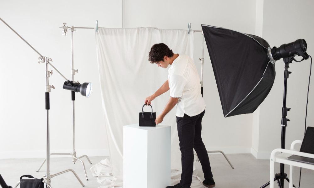 A person in a white short-sleeved shirt and black pant is adjusting a small black handbag placed on a white cube. The scene is a photography studio with a white fabric backdrop and various lighting equipment