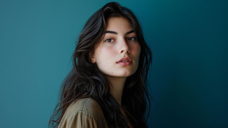 Portrait of a young woman with long dark hair, looking at the viewer against a dark teal background