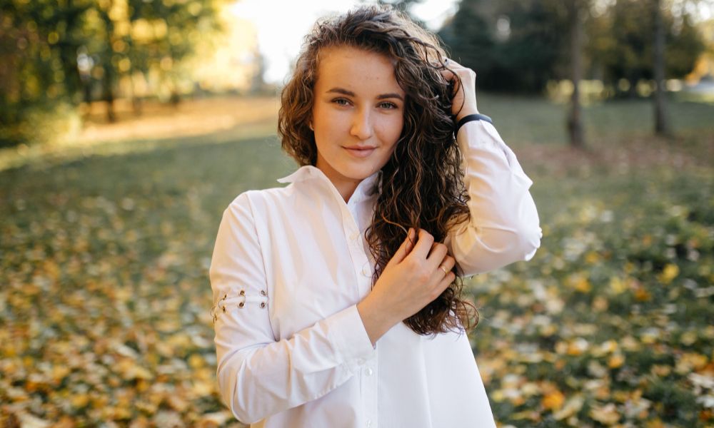 A smiling young woman with long, curly brown hair, wearing a white button-up shirt, poses in a natural setting with her hands near her hair, and a background of green grass and autumn leaves