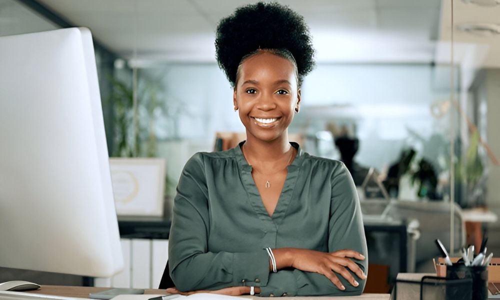 A young Black woman with an Afro hairstyle is smiling confidently at her desk in a modern office, her arms crossed, with a computer monitor to her right