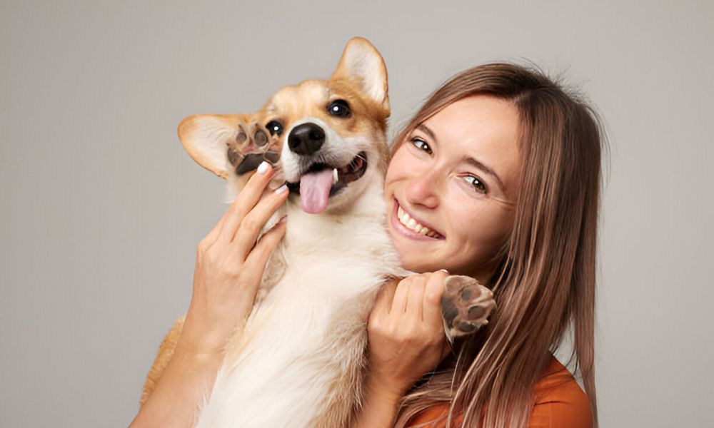 A cheerful woman with long brown hair smiles broadly while embracing a happy, panting corgi dog, both looking towards the viewer against a plain background