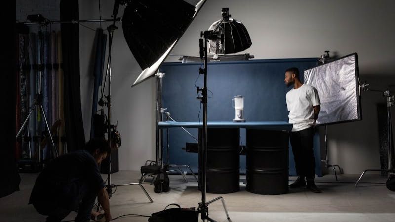 A photography studio scene with a man posing next to a blender on a table with a blue backdrop, while another person adjusts lighting equipment