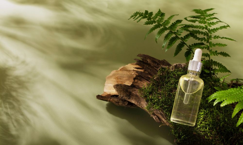 A clear dropper bottle filled with a light yellow liquid rests on a piece of moss-covered wood, accompanied by green fern leaves, against a muted green background with soft shadows