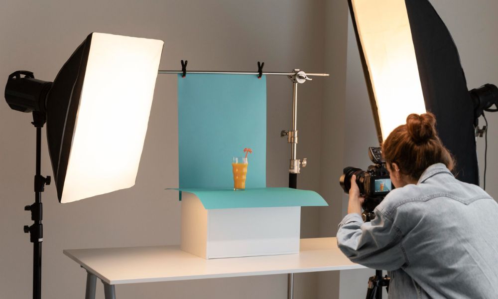 A female photographer is seen from behind, holding a camera and taking a photo of a glass of orange juice with a straw, set up on a white box with a light blue backdrop and surrounded by two large studio softbox lights