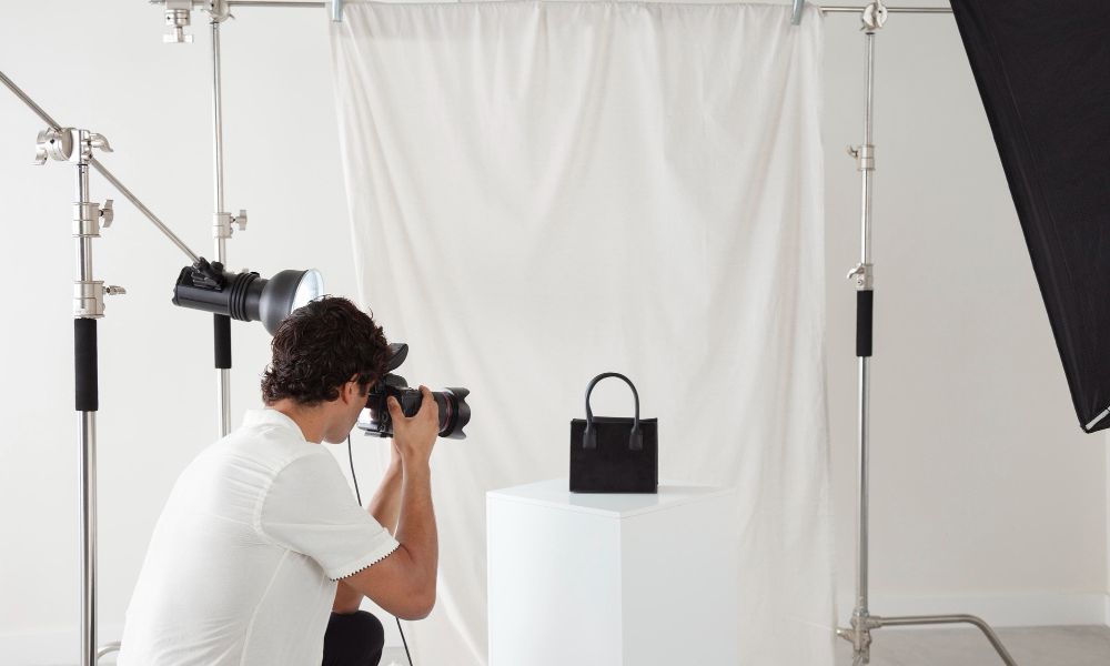 Photographer taking a picture of a black handbag in a professional studio setup with lights and a white backdrop