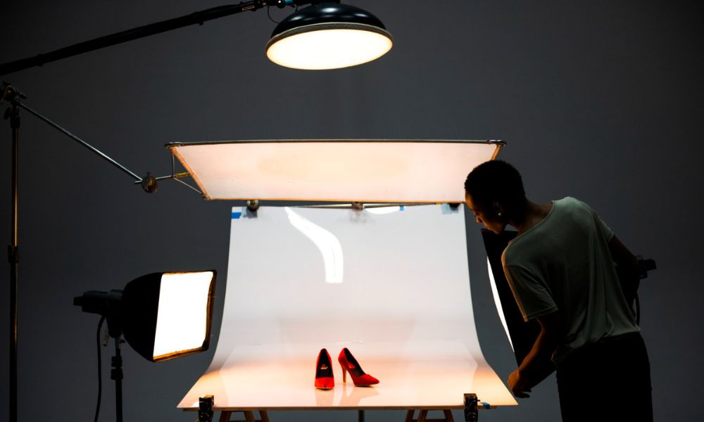 A professional product photography setup with multiple light sources in a dark studio. Two red high-heeled shoes are positioned on a seamless white backdrop, illuminated by various studio lights and diffusers, with a person adjusting equipment on the right
