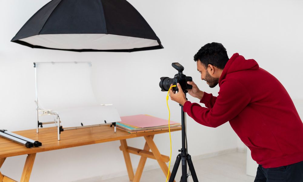 A male photographer in a red hoodie is adjusting the settings on a camera mounted on a tripod. In the background, there is a small white photo table with a seamless white backdrop, and a large black photography umbrella above