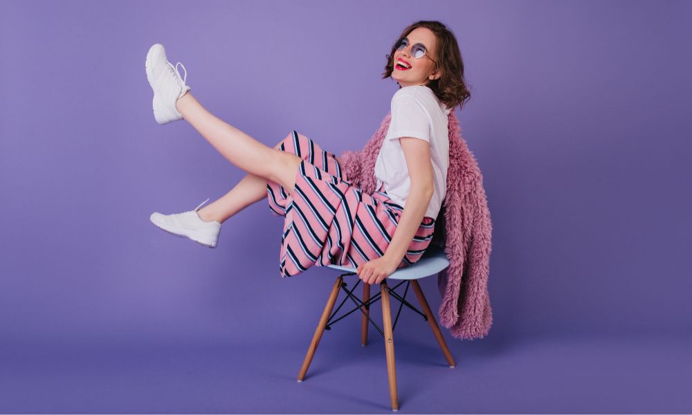 A cheerful woman in a striped skirt, white t-shirt, and white sneakers playfully poses on a chair against a purple background, with a pink fuzzy coat draped over her shoulders