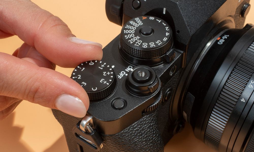 Close-up image of a person's hand adjusting the exposure compensation dial on a black mirrorless camera, showing detailed control dials and a zoom lens