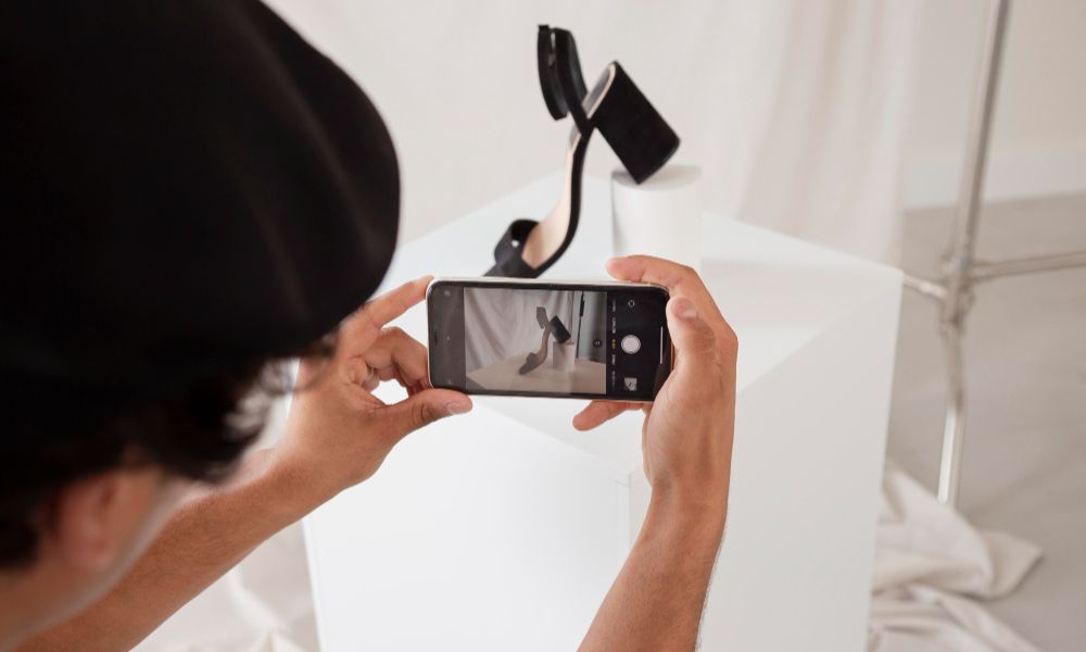 Person from behind, in a dark hat, photographing a black high-heeled shoe on white cubes with a smartphone