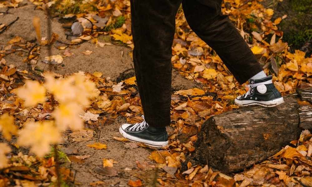A person wearing dark pants and black and white low-top sneakers is walking on a path covered in fallen orange and yellow autumn leaves. Part of a log or tree trunk is visible on the right
