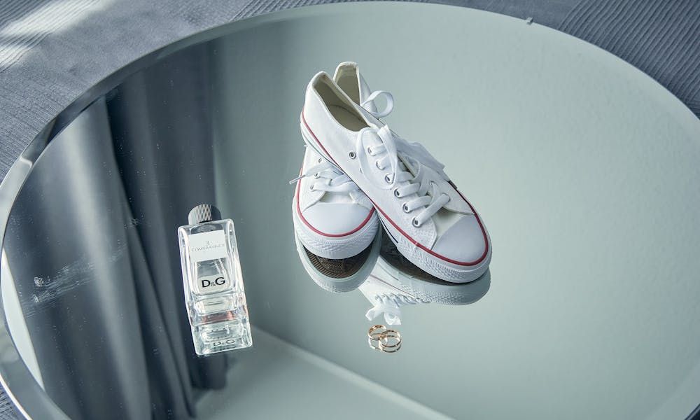 A pair of white sneakers with red trim and white laces, a bottle of D&G perfume, and two wedding rings are arranged on a circular mirrored surface, reflecting the items and the surrounding room