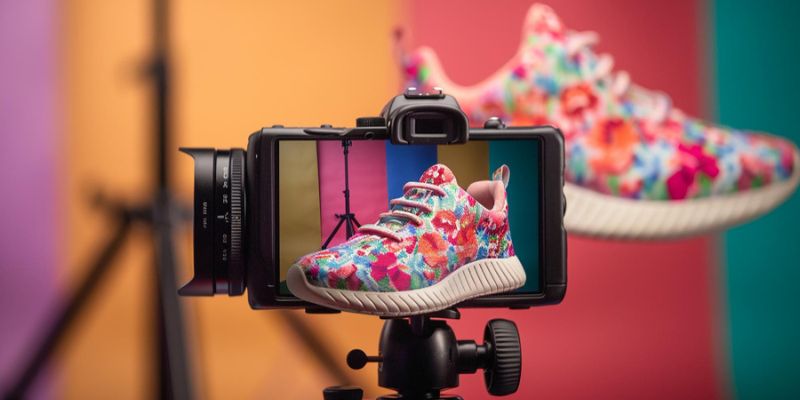 Camera on tripod showing a colorful floral sneaker on its screen, set against a multi-colored backdrop with another sneaker blurred in the background