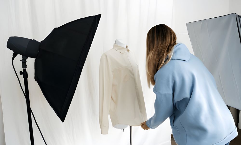 A female stylist, seen from behind, is adjusting a light-colored long-sleeve shirt on a white mannequin in a well-lit studio. Two large black studio lights flank the setup, and a white backdrop is visible