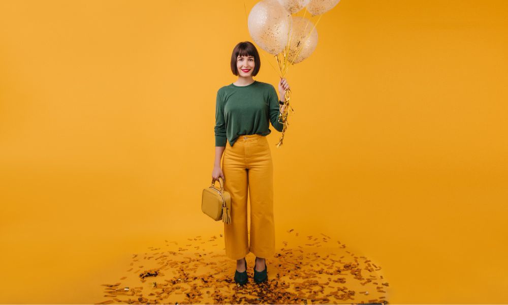 A smiling woman with a bob haircut and red lipstick stands on an orange background littered with confetti, holding a small yellow handbag and a bunch of clear balloons filled with golden confetti