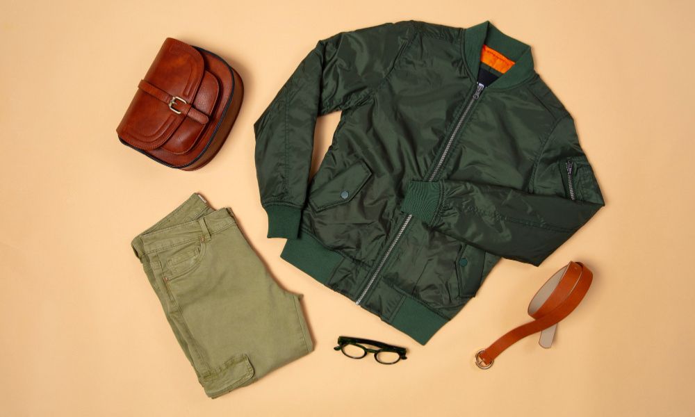 A flat lay of men's clothing and accessories on a light brown background. It features a dark green bomber jacket, olive green pants, a brown leather satchel, a brown leather belt, and black-framed eyeglasses